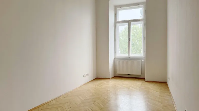 Helles, leeres Zimmer mit Fischgrätparkett und einem großen Fenster, das viel Tageslicht hereinlässt.