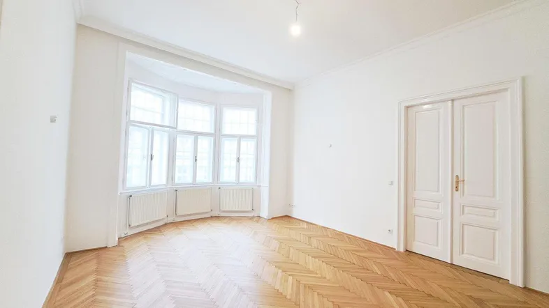 Heller, renovierter Raum mit klassischem Fischgrätparkett, Erkerfenstern und hohen Doppeltüren.