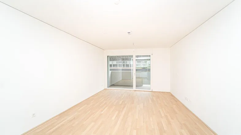 Helles, leeres Zimmer mit Parkettboden und Zugang zu einem Balkon, ideal als Wohnbereich.
