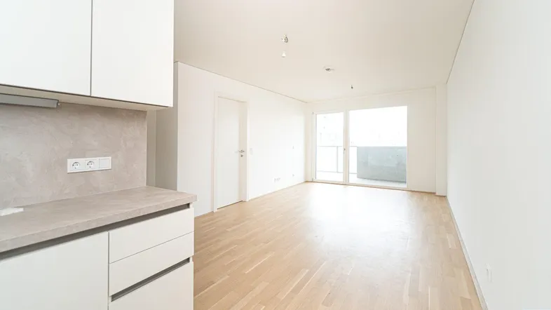 Heller Wohnbereich mit integrierter Küchenzeile, Parkettboden und Zugang zum Balkon.