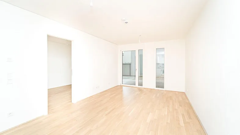 Helles, leeres Zimmer mit Holzboden und Zugang zu einem Außenbereich durch Glastüren.