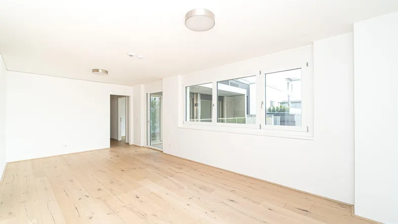 Helles, leeres Zimmer mit modernem Holzboden und großen Fenstern, die viel Tageslicht hereinlassen.