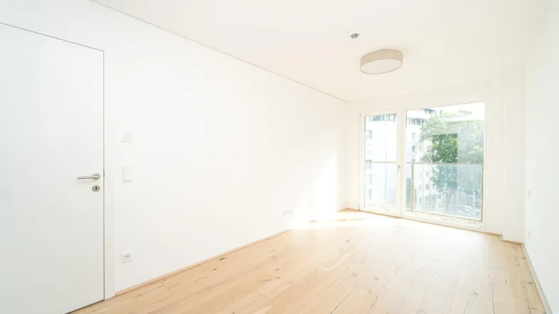 Helles, unmöbliertes Zimmer mit Holzboden und Zugang zu einem französischen Balkon.