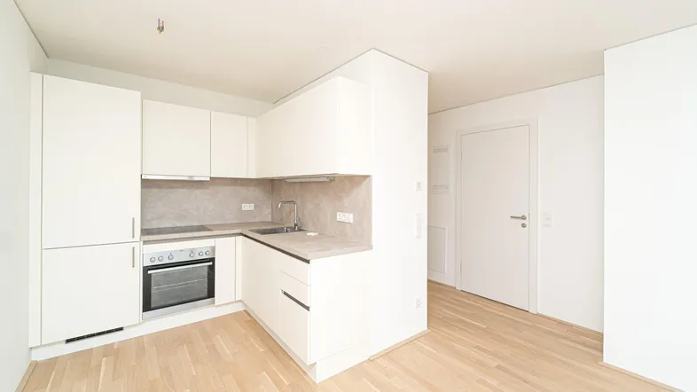 Moderne, kompakte Einbauküche mit weißen Schränken, integriertem Backofen und hellem Parkettboden.
