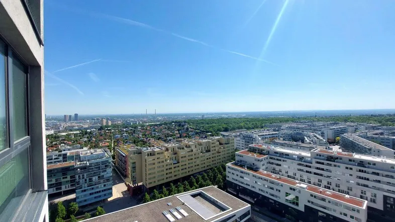 Panoramablick über die Stadt mit zahlreichen Gebäuden und weiten Grünflächen am Horizont unter blauem Himmel.