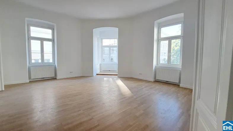 Helles Zimmer mit Parkettboden, hohen Fenstern und einem Durchgang zu einem weiteren Raum.