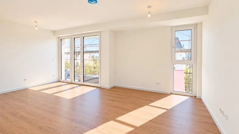 Helles, leeres Wohnzimmer mit großen Fenstern, Holzboden und viel natürlichem Licht.