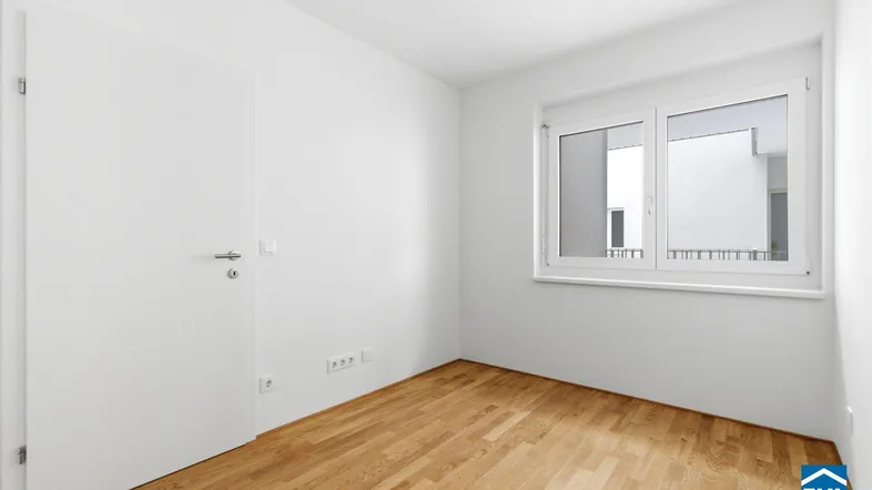 Helles, leeres Zimmer mit Parkettboden und einem Fenster, das Tageslicht hereinlässt.