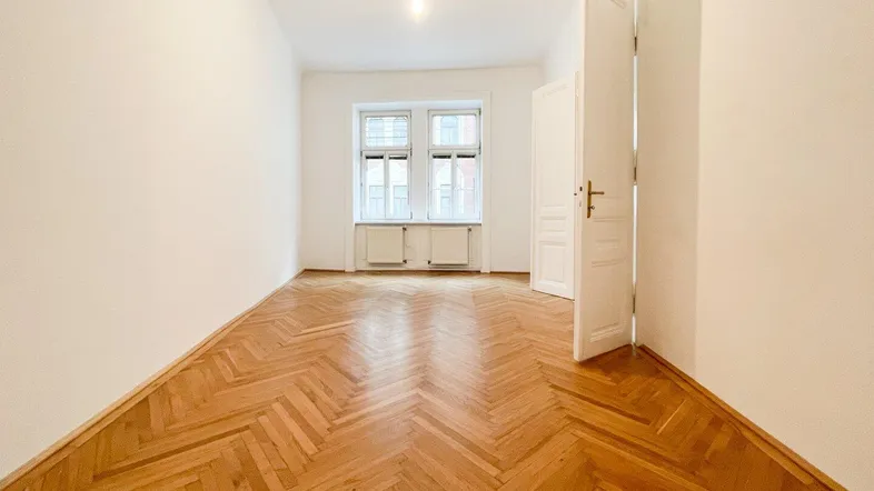 Helles Zimmer mit Fischgrätparkettboden, weißen Wänden und großen Fenstern.