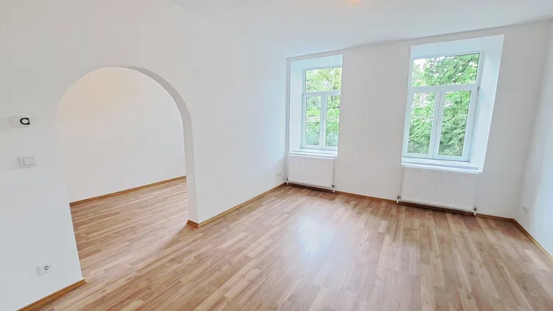 Heller, leerer Raum mit Laminatboden, zwei Fenstern und einem Rundbogen zu einem weiteren Bereich.