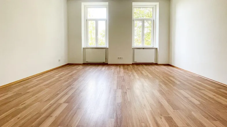 Heller, leerer Raum mit Laminatboden, weißen Wänden und zwei großen Fenstern mit Blick ins Grüne.