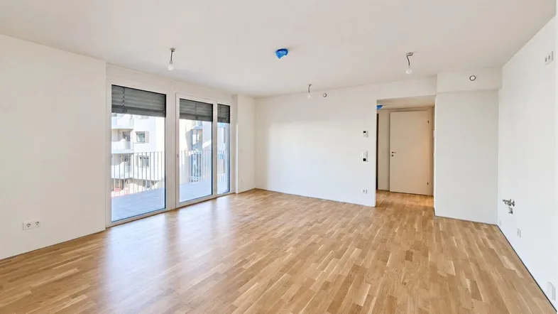 Heller, leerer Wohnraum mit Parkettboden, großen Fenstern und direktem Zugang zum Balkon.