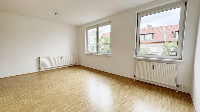 Helles, leeres Zimmer mit Parkettboden und zwei Fenstern, die viel Tageslicht hereinlassen.