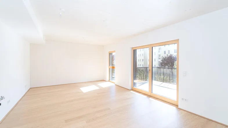 Helles, leeres Wohnzimmer mit hellem Holzfußboden und großzügigem Zugang zum Balkon.