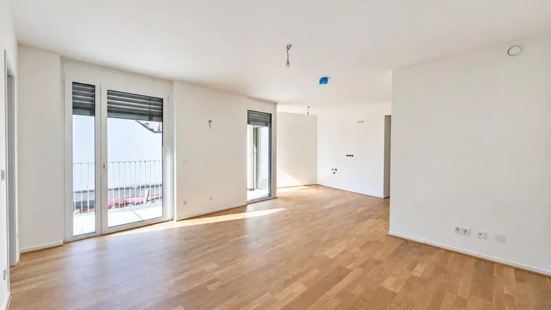Helles, leeres Wohnzimmer mit Parkettboden und Zugang zu einem Balkon in einem Neubau.