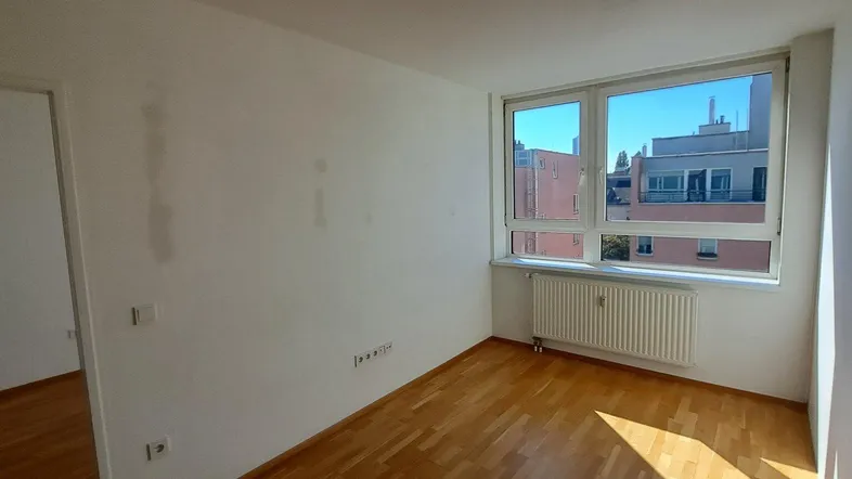 Helles, leeres Zimmer mit Parkettboden, großem Fenster und Blick auf die Stadt.
