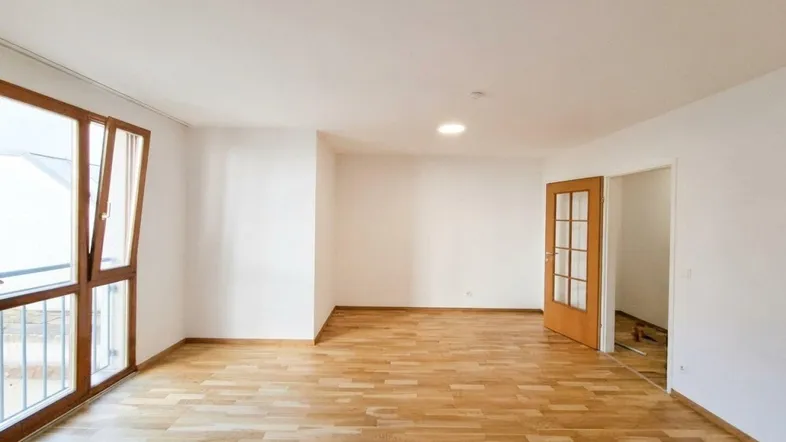 Helles, leeres Wohnzimmer mit Holzboden, großen Fenstern und Zugang zu einem Balkon.