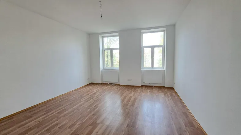 Helles, leeres Zimmer mit Laminatboden und zwei großen Fenstern, bereit zur individuellen Gestaltung.