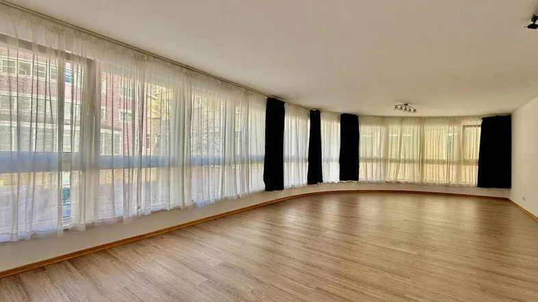 Geräumiges Wohnzimmer mit großen, gebogenen Fenstern und hellem Laminatboden, das viel Tageslicht hereinlässt.