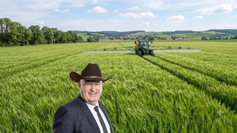 Ein Mann im Anzug mit Cowboyhut steht vor einem grünen Feld, auf dem ein Traktor spritzt.
