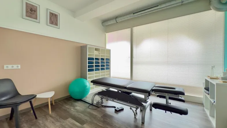 Heller Behandlungsraum mit Therapieliege, Sportgeräten und großen Fenstern mit Jalousien.