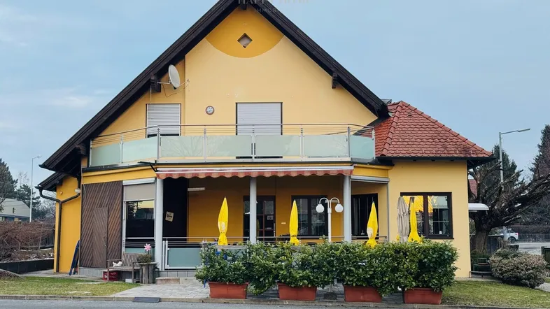 Gelbes Gebäude mit rotem Ziegeldach, Balkon und überdachtem Außenbereich mit Sonnenschirmen.