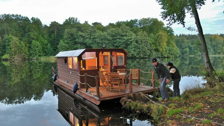 Gemütliches Hausboot auf einem See, mit beleuchteter Terrasse und Sitzgelegenheiten am Ufer.