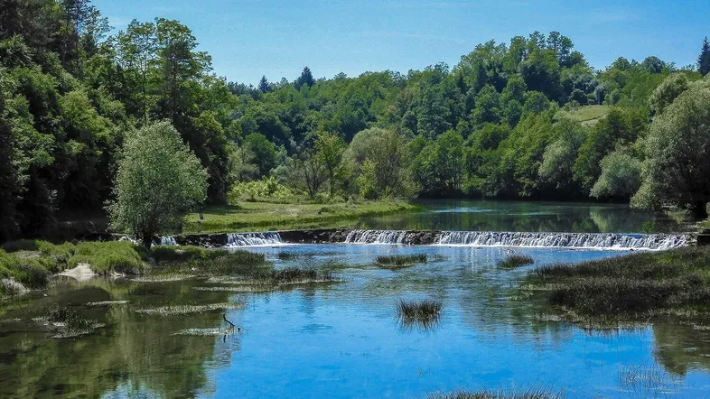 Flusslandschaft mit kleinem Wasserfall, umgeben von dichtem Grün und blauem Himmel.