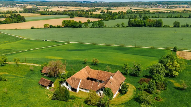 Luftaufnahme eines weitläufigen Bauernhofs mit mehreren Gebäuden, umgeben von grünen Feldern und Wäldern unter blauem Himmel.