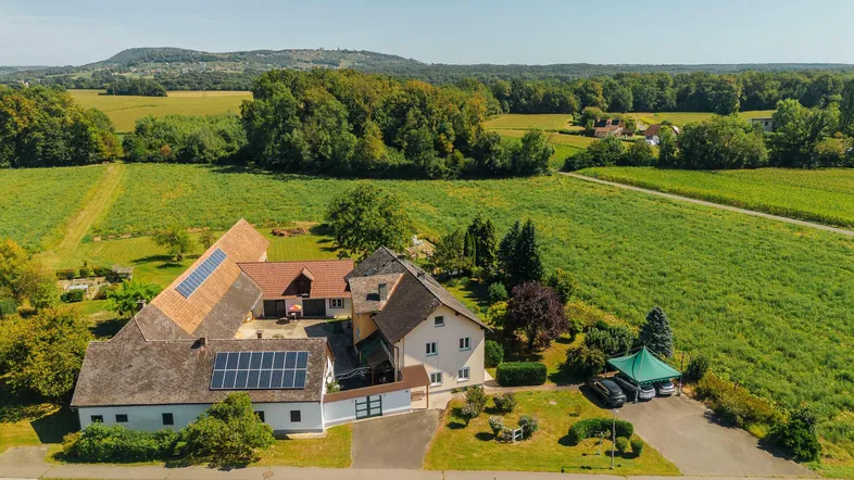 Luftaufnahme eines Anwesens mit mehreren Gebäuden, umgeben von grünen Feldern und Wäldern, mit Solaranlagen auf dem Dach.