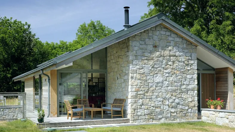 Moderne Steinfassade mit großer Terrasse und Sitzgelegenheiten im Grünen, umgeben von Bäumen.