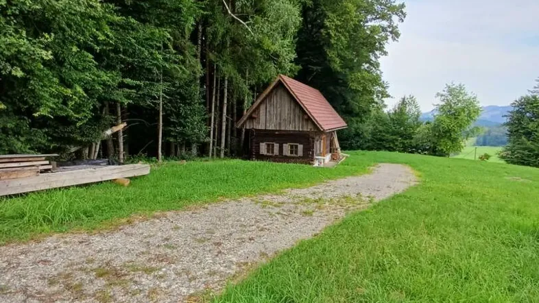 Rustikale Holzhütte mit rotem Dach am Waldrand, umgeben von grünen Wiesen und einem Schotterweg.