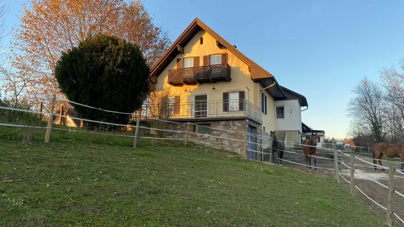 Ländliches Haus mit Satteldach, Balkonen und angrenzender Pferdekoppel bei Sonnenuntergang.