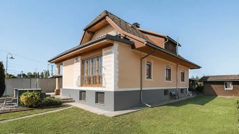 Seitenansicht des Hauses mit Balkon, gepflegtem Rasen und ländlicher Umgebung an einem sonnigen Tag.