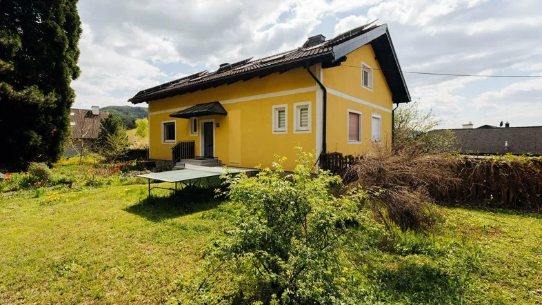 Gelbes Einfamilienhaus mit Satteldach und großem Gartenbereich unter bewölktem Himmel.