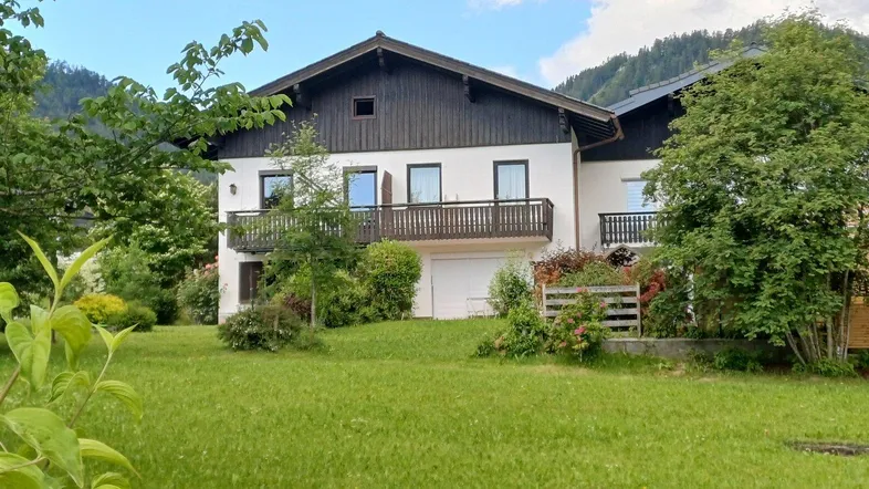 Hausfassade mit Balkon und gepflegtem Garten, umgeben von grüner Natur und Bergen.