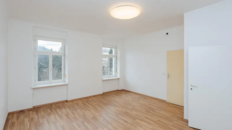 Helles Zimmer mit Parkettboden, weißen Wänden und zwei Fenstern für viel Tageslicht.