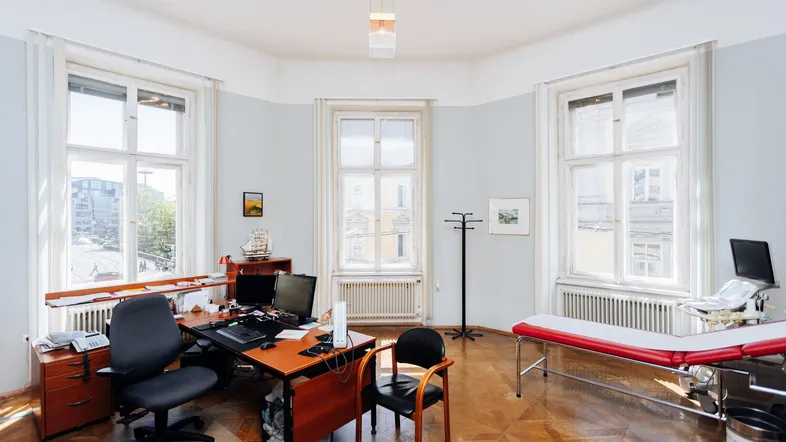 Geräumiges Arztzimmer mit Schreibtisch, Untersuchungsliege und großen Fenstern, die viel Tageslicht hereinlassen.
