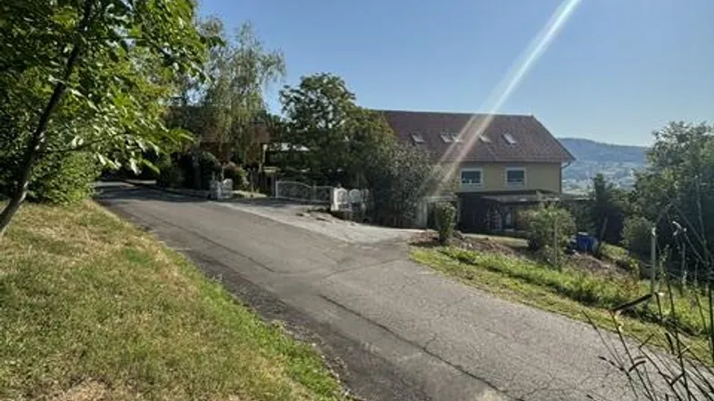 Blick auf ein Wohnhaus am Hang mit sonniger Umgebung und einer Zufahrtsstraße.