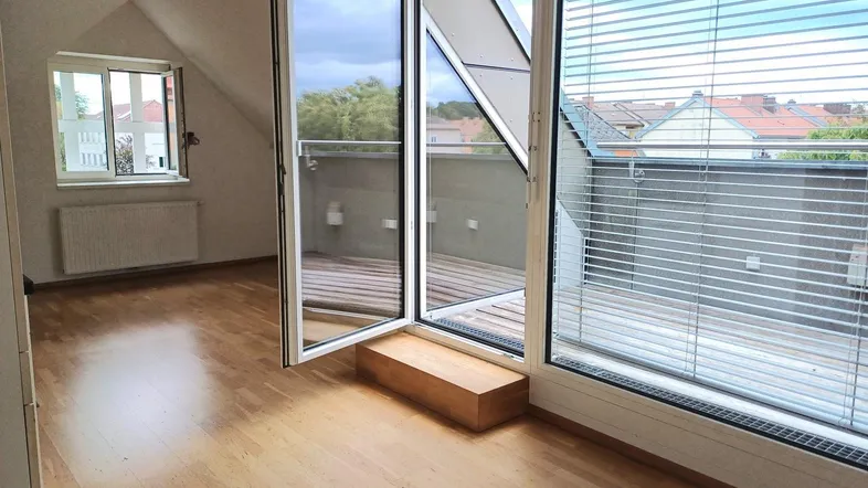 Helles Zimmer im Dachgeschoss mit Holzboden, Heizkörper und Zugang zu einer Terrasse mit Blick ins Grüne.