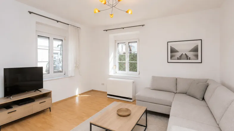 Helles Wohnzimmer mit modernem Sofa, Holzboden und zwei Fenstern, die viel Tageslicht hereinlassen.