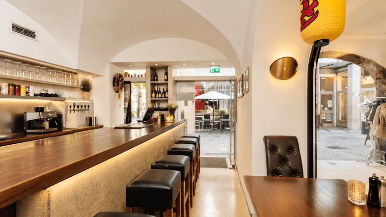 Heller Restaurantbereich mit langer Holztheke, Barhockern und gewölbten Decken, führt zu einem Außenbereich.