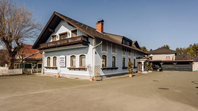 Traditionelles Gebäude mit hellblauer Fassade, dunklem Dach und einem Balkon, umgeben von einem großen Vorplatz.