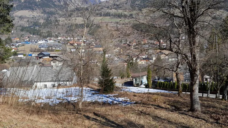 Panoramablick auf ein Dorf im Tal mit schneebedeckten Bergen im Hintergrund und Bäumen im Vordergrund.