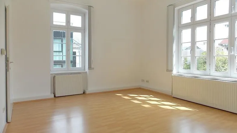 Helles, leeres Wohnzimmer mit Laminatboden, weißen Wänden und großen Fenstern.