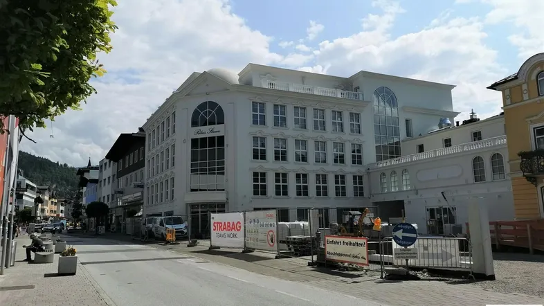 Weißes Gebäude mit klassischer Architektur und vielen Fenstern, umgeben von einer städtischen Straße und Baustellenabsperrungen.