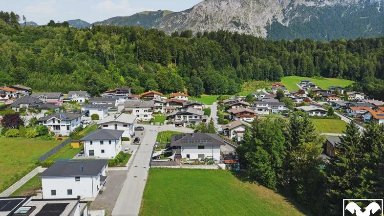 Luftaufnahme einer Wohnsiedlung mit Häusern, grünen Wiesen und Bergen im Hintergrund.