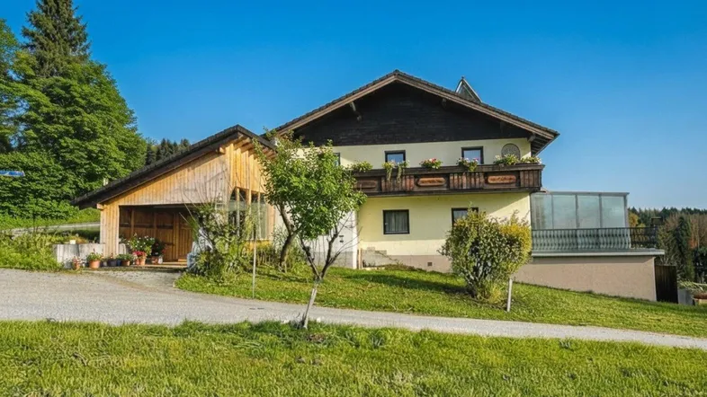 Traditionelles Haus mit Holzbalkon, Wintergarten und Carport in ländlicher Umgebung, umgeben von viel Grün.