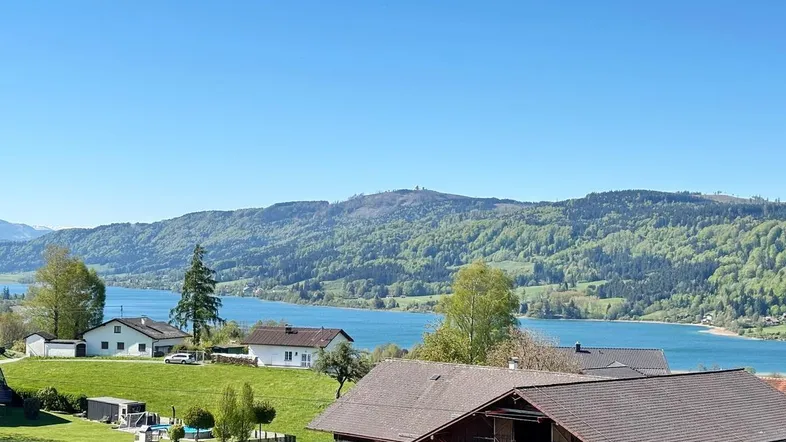 Panoramablick auf einen See und die umliegende grüne Landschaft mit vereinzelten Häusern unter blauem Himmel.