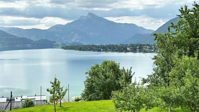 Panoramablick auf den See und die umliegenden Berge mit grüner Landschaft im Vordergrund.